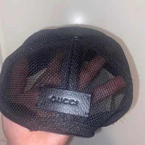 Äkta svart Gucci keps  - Säljer en svart keps från Gucci med klassiskt GG-mönster framtill och mesh-paneler bak. Kepsen har en böjd skärm och justerbar baksida i läder med präglad logotyp. Materialet är en mix av polyester och läderdetaljer. Perfekt för dig som gillar streetwear och designeraccessoarer.