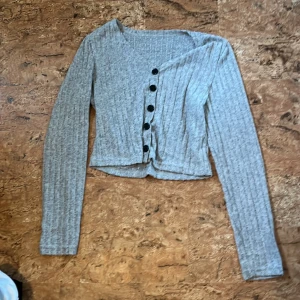 Grå ribbad långärmad kofta M - Grå ribbad långärmad kofta med svarta knappar framtill. Koftan har en croppad passform och är tillverkad i mjuk polyesterblandning. Perfekt för dig som gillar en enkel men ändå trendig look.