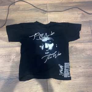Pop Smoke tröja från Vlone - Svart t-shirt från Vlone med stor vit V i rökdesign på ryggen och vit text samt grafiskt tryck framtill. Klassisk passform med rund hals och korta ärmar. Perfekt för dig som gillar streetwear och vill sticka ut med en unik design.