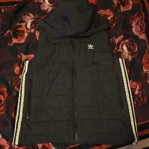 Svart dunväst med huva från Adidas - Svart dunväst från Adidas med huva och klassiska vita ränder längs sidorna. Västen har dragkedja framtill, två fickor med dragkedja och Adidas-logga på bröstet. Perfekt för lager-på-lager och streetstyle.
