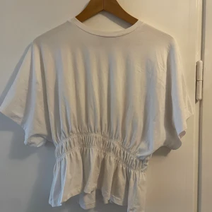 Vit topp med resår i midjan - Säljer en vit topp med korta ärmar och rund hals. Toppen har resår i midjan som ger en snygg rynkad effekt och volang nedtill. Tillverkad i mjuk bomull. Den har en liten sminkfläck vid kragen, går nog att tvätta bort har ej testat vanish osv. 