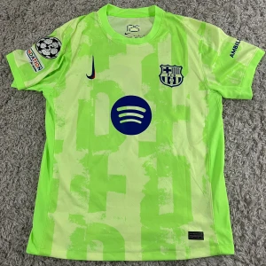 FC Barcelona Lamine Yamal Nike tröja M - Säljer en FC Barcelona matchtröja med Lamine Yamal 19 tryck. Tröjan är grön med unikt mönster, klubbmärke och Nike-logga på bröstet samt Spotify-logga i mitten. Korta ärmar, Dri-FIT-material och Champions League patch på ärmen. Perfekt för dig som älskar Barca. Den passade inte mig, därför säljer jag den.