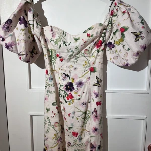 Blommig kortklänning från H&M - Superfin kortklänning från H&M i ljus bas med färgglatt blommönster, puffiga ärmar och snörning i ryggen. Klänningen har en sweetheart-ringning och är gjord i en linnemix. Storlek 34, i fint skick.