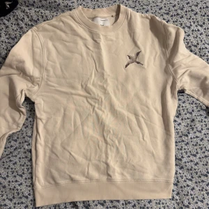 Axel Arigato-Gräddvit Sweatshirt - Gräddvit sweatshirt med rund hals och ribbade muddar. På bröstet finns en broderad fågel i grå toner som ger en stilren detalj. Perfekt för dig som gillar enkel men snygg design. Passar till jeans eller mjukisbyxor.