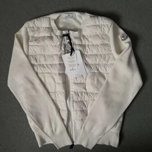 Vit quiltad jacka från Moncler - Exklusiv vit jacka från Moncler med quiltad front och ärmar i mjuk stickad kvalitet. Tröjan har dragkedja framtill, rund halsringning och Moncler-logga på ärmen. Kvitto och dustbag ingår.