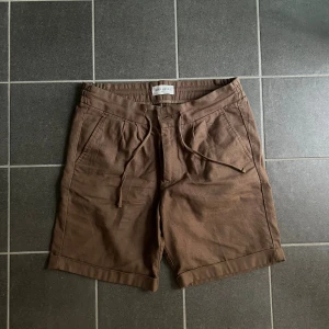 Bruna linneshorts från John Henric - Säljer ett par bruna shorts från John Henric med snörning i midjan och avslappnad passform. Gjorda i en skön mix av bomull och linne, perfekta för varma dagar. Shortsen har fickor fram och bak samt uppvikta benslut för en chill look.