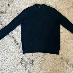 Mörkblå stickad tröja från H&M - Mörkblå stickad tröja från H&M i storlek M. Tröjan har rund halsringning, ribbade muddar och ett diskret stickat mönster. Perfekt för lager-på-lager och passar till jeans eller chinos.