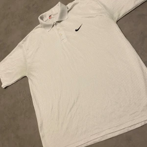 Vintage baggy Nike Airliner piké  - Vit pikétröja från Nike Airliner med diskret ribbat mönster och svart swoosh-logga på bröstet. Klassisk krage och knäppning med tre knappar. Perfekt för en clean och sportig stil.