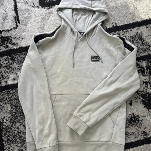 Ea7 hoodie - Snygg ljusgrå hoodie från EA7 Emporio Armani med svart logotyp på bröstet och svarta detaljer på axlarna. Tröjan har huva med snörning, stor magficka och långa ärmar. Perfekt för chill eller streetwear. Materialet är mjuk bomull med insida i fleece.