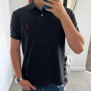 Svart Polo Ralph Lauren piké M - Svart t-shirt från Polo Ralph Lauren i klassisk pikéstil med krage och två knappar. Röd broderad logga på bröstet. Tillverkad i mjuk bomull som är skön mot huden. Passformen är normal och ärmarna är korta. Perfekt för en clean och stilren look. Modellen är 177 68kg och sitter bra, skriv gärna vid frågor eller funderingar. Priset är ej hugget i sten