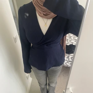 Mörkblå omlottopp med v-ringning - Snygg marinblå omlottopp med v-ringning, långa ärmar med rosett på baksidan. Toppen har en figurnära passform och markerad midja som ger en stilren siluett. 