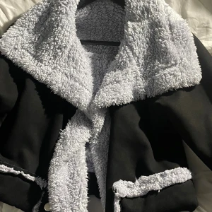 Svart teddyjacka med grå fuskpäls - Säljer en svart teddyjacka med breda grå fuskpälsdetaljer längs krage, ärmslut och fickor. Jackan har en oversized känsla och är fodrad med mjuk teddy på insidan. Perfekt för kalla dagar när du vill vara både varm och snygg.