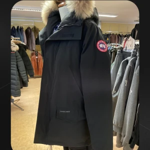 Svart dunjacka från Canada Goose L - Säljer en svart Canada Goose dunjacka i storlek L med klassisk patch på ärmen och stor huva med fluffig päls. Jackan har dragkedja och stora fickor framtill, perfekt för kalla vinterdagar. Jackan är storlek Large men sitter mer som Medium. Inga tecken på användning.