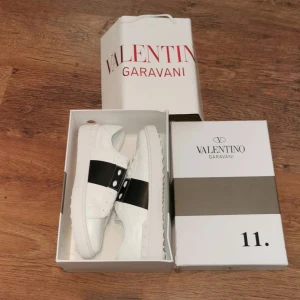Valentino Garavani Open sneakers vit/svart - Snygga Valentino Garavani Open sneakers i vitt skinn med bred svart läderdetalj över vristen. Klassisk rund tå, vita snören och ikoniska nitar baktill på sulan. Perfekta för dig som vill sticka ut med lyxig streetstyle.