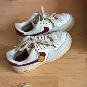 Nike Air Force 1 vit/röd sneakers - Nike Air Force 1 sneakers i vitt skinn med röda detaljer och swoosh. Klassisk låg modell med perforerad tå, beige sula och guldfärgade detaljer på snörningen. Snygg retrostil som passar perfekt till streetwear.