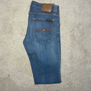 Nudie Jeans - Säljer ett par klassiska blå jeans från Nudie Jeans med snygga slitningar och kontrastsömmar. Jeansen har dragkedjegylf, fem fickor och Nudie-logga på bakfickan. Perfekt passform och riktigt sköna att bära. Modellen är tight long jonh 