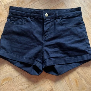 Svarta jeansshorts från H&M, strl 34 - Svarta jeansshorts från H&M i storlek 34. Klassisk femficksmodell med hög midja, gylf med dragkedja och knapp i guld. Shortsens ben är uppvikta och har bälteshällor. Perfekta för sommarens alla dagar.