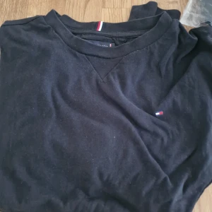 Svart långärmad tröja Tommy Hilfiger stl:164 - Svart långärmad tröja från Tommy Hilfiger i mjuk bomull. Klassisk design med liten broderad logga på bröstet och Hilfigers signaturrand på insidan av kragen. Perfekt basic-plagg med normal passform och rund hals.