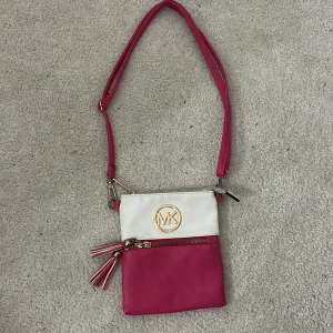 Rosa och vit axelväska från Michael Kors - Säljer en snygg axelväska från Michael Kors i rosa och vitt skinn. Väskan har justerbar axelrem, guldfärgade detaljer, dragkedja och dekorativa tofsar. Framsidan pryds av MK-logga i guld. Perfekt storlek för mobilen och det viktigaste.