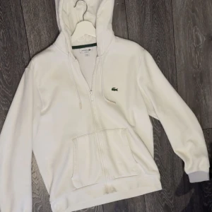 Vit hoodie från Lacoste med dragkedja storlek M - Snygg vit hoodie från Lacoste Storlek L men passar bättre på M, väldigt bra skick.