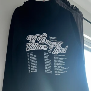 Svart Hov1 hoodie med turnétryck - Svart hoodie från Hov1 med stort vitt turnétryck på ryggen där det står 'Vi Dansar Vidare I Livet' och turnédatum. Klassisk huva och avslappnad passform, perfekt för dig som gillar svensk musik och streetstyle.