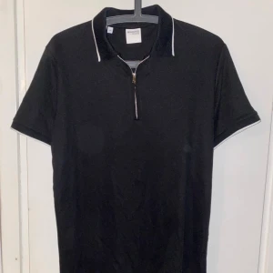 Svart Selected Homme Halv zip - Svart t-shirt från Selected Homme med vit kant på krage och ärmslut. Klassisk pikékrage och snygg dragkedja framtill. Perfekt för dig som gillar stilren och enkel design.