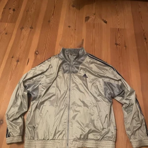 Silvrig vindjacka från Adidas - Silvrig vindjacka från Adidas med klassiska svarta ränder längs ärmarna och mesh-detaljer vid axlar och armveck. Jackan har dragkedja framtill, ståkrage och två fickor. Perfekt för dig som gillar sportig och clean stil.