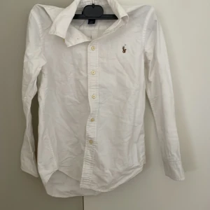Vit skjorta från Ralph Lauren - Klassisk vit skjorta från Ralph Lauren med broderad logga på bröstet. Skjortan har lång ärm, knappar framtill och är tillverkad i bomull. Snygg passform och tidlös design som funkar till allt.