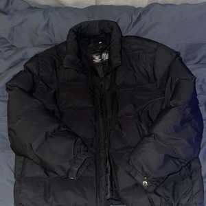 Svart pufferjacka med dragkedja - Säljer en svart pufferjacka från H&M med hög krage och dragkedja framtill. Jackan har långa ärmar, två sidofickor samt en innerficka och en ficka på bröstet med dragkedja. Perfekt för kalla dagar när du vill hålla dig varm och ändå se chill ut. Litet hål på högerarm men knappt så att det syns. Kan absolut sänka priset vid snabb affär