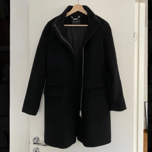 Svart kappa från STOCKH LM - Stilren svart kappa från STOCKH LM i ullblandning 50%ull.  Rak modell med hög krage, långa ärmar och två fickor med lock framtill. Stängs med dragkedja och har ett klassiskt, minimalistiskt uttryck som passar perfekt till höst och vinter. Varm o skön, knappt använd, som ny!  Nypris 2999kr