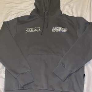 Grå hoodie från Hoodrich med tryck - Snygg grå hoodie från Hoodrich med vit och svart texttryck både fram och bak. Klassisk känguruficka, huva med snörning och streetwear-vibe. Perfekt för dig som gillar stilrena och trendiga plagg med tydlig logga.