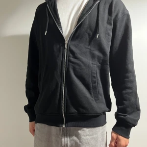 Svart hoodie med dragkedja från H&M - Svart hoodie från H&M i loose fit med dragkedja framtill och snörning i huvan. Tröjan har långa ärmar, två fickor på sidorna och är tillverkad i mjuk bomullsmix. Perfekt för en chill och avslappnad stil.