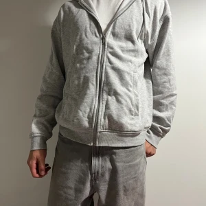 Grå hoodie från H&M loose fit - Säljer en grå hoodie från H&M i loose fit med dragkedja framtill och huva med snören. Tröjan har långa ärmar, ribbade muddar och två fickor på sidorna. Perfekt för chill dagar och enkel att matcha med jeans eller mjukisbyxor.