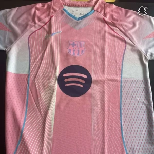 Super fin limited edition Lamine Yamal 10 - Barcelona fotbollströja i rosa och ljusblått från Nike, med klubbmärke och Spotify-logga på bröstet. Tröjan har korta ärmar, rund hals med ljusblå kant och tunna vita linjer i mönstret. Tillverkad i lätt och ventilerande funktionsmaterial. Det är storlek M och passar perfekt på kroppen 