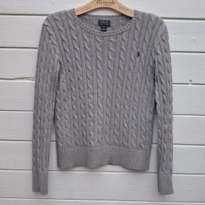 Grå kabelstickad damtröja Ralph Lauren - Superfin Ralph Lauren kabelstickad tröja, grå med lila logga. Märkt stl XL(16), sitter ungefär som en vuxen XS/S, se mått. Toppenskick, inga fläckar eller hål, mjuk och stretchig kvalitet.  Mått: Armhåla till armhåla 45cm, bredd längst ner 42cm, längd från mitt på axeln 53cm.