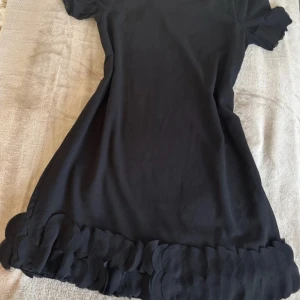 Svart kortklänning med volangdetaljer - Supersöt svart kortklänning med rund hals och korta ärmar. Klänningen har unika volangdetaljer i form av cirklar längs fållen och vid ärmsluten, vilket ger en cool och lekfull look. Perfekt för dig som gillar stilrena plagg med en twist.