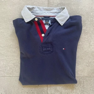 Marinblå pikétröja Tommy Hilfiger - Snygg marinblå pikétröja från Tommy Hilfiger. Tröjan har korta ärmar, klassisk krage med ljusblåa ränder och knappar framtill. Detaljer i rött och mörkblått vid knäppningen samt broderad logga på bröstet. Perfekt för en clean och stilren look.