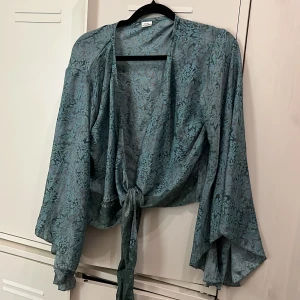 Blommig blå silkesblus med knyt - Säljer en tunn och flowy blus i 100% silke med blått blommönster. Blusen har vida ärmar och knyt framtill för en avslappnad och cool look. Perfekt för dig som gillar unika detaljer och mjuka material.