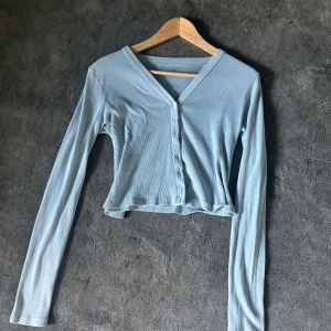 Ljusblå ribbad kofta från brandy Melville - Säljer en ljusblå ribbad blus från Brandy melville med croppad passform. Toppen har långa ärmar, v-ringning och knäppning framtill med små vita knappar. 
