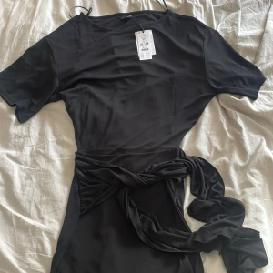 Svart kort klänning med knyt - Supersnygg svart kort klänning från Gina Tricot i slätt material. Klänningen har korta ärmar och ett brett knytband i midjan som ger en cool och trendig look. Perfekt för dig som gillar stilrena och enkla plagg med en twist. Prislapp är kvar så har aldrig använt.