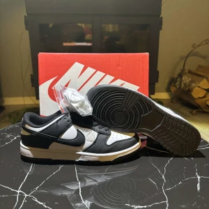 Nike Dunk Low svart/vit storlek 39 - Nike Dunk Low sneakers i svart och vitt med klassisk Swoosh på sidorna. Skorna har snörning, perforerad tå och platt sula. Tillverkade i skinn och syntetmaterial. Levereras med extra vita skosnören. Perfekt för dig som gillar streetstyle.