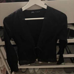 Svart bolerotopp med broderi från Zara - Svart bolerotopp från Zara med långa ärmar, knytning framtill och vackra broderade detaljer längs nederkanten. Tunn och lätt i materialet, perfekt att slänga över ett linne eller klänning. Snygg v-ringning och diskret spetskant för extra detalj.