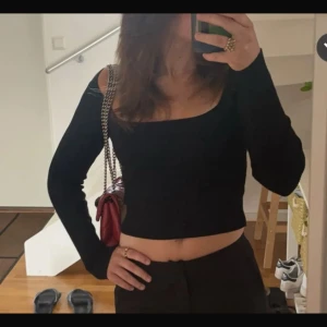 Svart ribbad croppad topp Bershka - Säljer en svart ribbad croppad topp från Bershka med lång ärm och fyrkantig urringning. Toppen har en unik detalj med öppen axel och hög halsringning. Perfekt för dig som gillar trendiga och stilrena plagg. Den har ett litet trasig söm i fram vid ringningen men som går att laga 