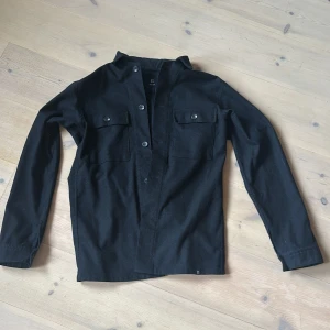 Svart overshirt från Boggi Milano - Stilren svart overshirt från Boggi Milano i mjukt material. Skjortan har två bröstfickor med knappar, klassisk krage och långa ärmar. Perfekt att slänga över en t-shirt för en clean look.