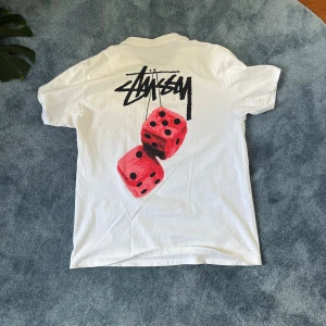 Vit Stüssy t-shirt med tärningar - Exklusiv Vit t-shirt från Stüssy med klassisk logga på bröstet och stor grafisk print med röda tärningar på ryggen. Skön passform och tillverkad i mjuk bomull. Perfekt för dig som gillar streetwear och vill sticka ut lite extra. Nypris 1300