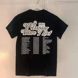 Svart Hov1 Stockholm t-shirt - Svart t-shirt från Hov1 med vit text 'Hov1 STOCKHOLM 2015 - 2025' på framsidan och turnédatum samt platser på baksidan. Klassisk rund hals och korta ärmar. Perfekt för dig som älskar svensk musik och vill visa din support för Hov1. Du får den helt oöppnad och ny i plastförpackningen!