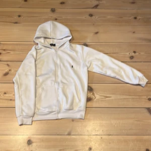 Vit hoodie från Polo Ralph Lauren S/M - Snygg vit hoodie från Polo Ralph Lauren med dragkedja och klassisk broderad logga på bröstet. Tröjan har huva, känguruficka och är tillverkad i mjuk bomull. Perfekt för en chill och stilren look.