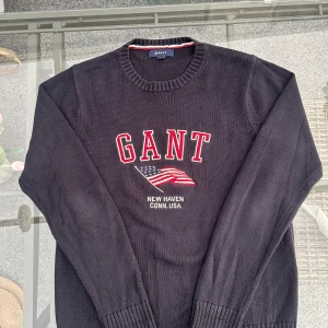 GANT stickad tröja navy S – USA flagga & logga - 🔥 Preppy classic från GANT – marinblå stickad tröja i storlek S. Fronten pryds av ikoniska GANT-loggan och amerikanska flaggan, med den klassiska New Haven Conn. USA texten.  ✅ 100% bomull – skön & slitstark ✅ Perfekt oversized på XS / true to size på S ✅ College-vibbar, preppy men funkar lika bra i street-styling  Matcha med jeans, chinos eller shorts för en clean och tidlös look. Tröjan är i mycket fint skick, redo för nästa ägare.