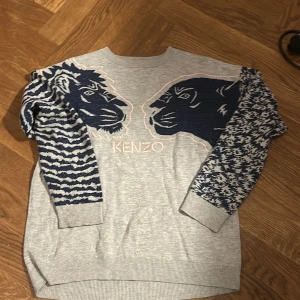 Grå Kenzo tröja med blå mönster - Säljer en grå tröja från Kenzo med stora blå lejonmotiv framtill och coola mönstrade ärmar i blått och vitt. Tröjan har rund hals och ribbade muddar. 