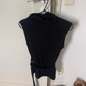 Svart ribbad polotopp med bälte - Säljer en svart ribbad polotopp utan ärmar. Toppen har en hög krage och ett matchande bälte i midjan som ger en snygg siluett. Perfekt att styla med jeans eller kjol för en clean look.